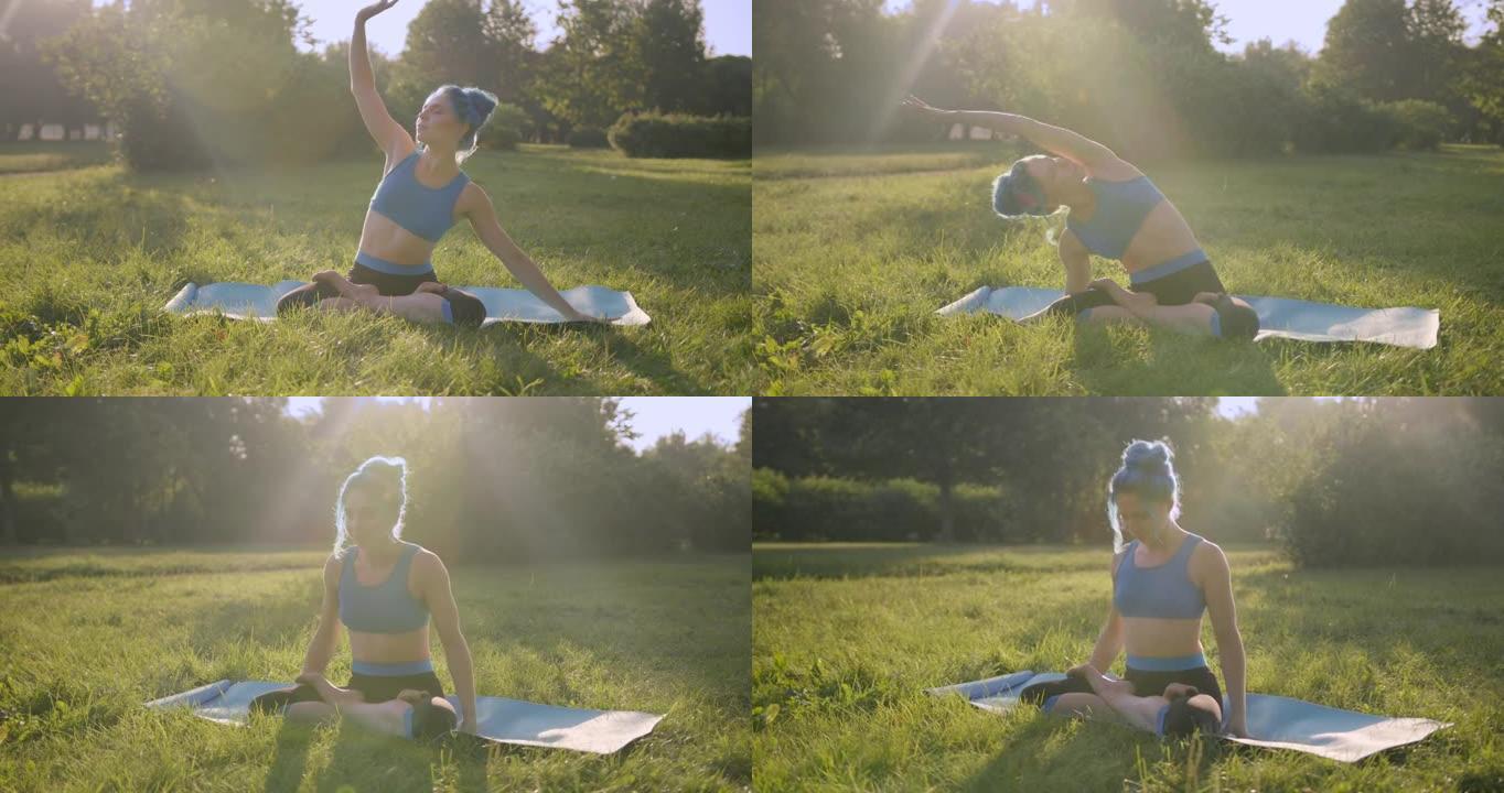 迷人的年轻女子，蓝色的头发，穿着蓝色的瑜伽服装，在公园的草地上做瑜伽，在日出或日落时，左右伸展，在瑜