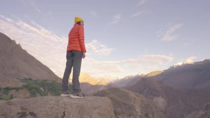 后视图，自由快乐的旅行者年轻女性穿着橙色的冬季夹克站在山顶上，在日出或日落时可以看到风景。体验式旅行