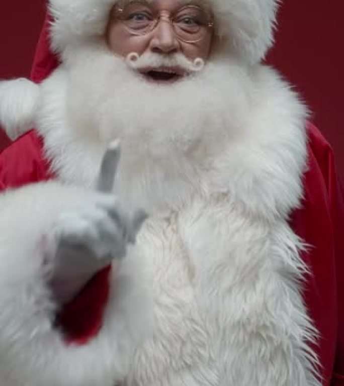 红色背景上的圣诞老人的垂直视频环顾四周，向我们展示了一个沉默的标志，将他的手指笑着放在嘴唇上，