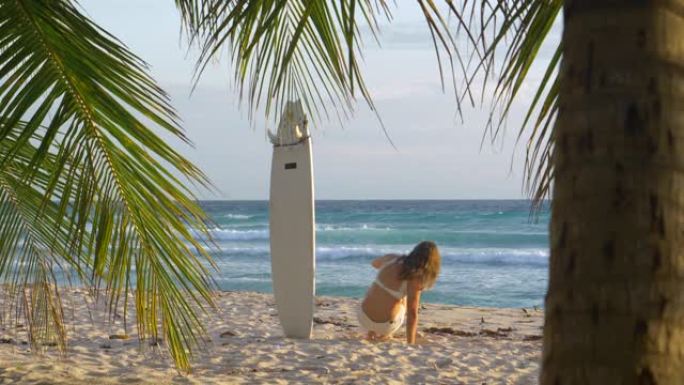 慢动作女子将冲浪板粘在沙滩上坐下观察日出