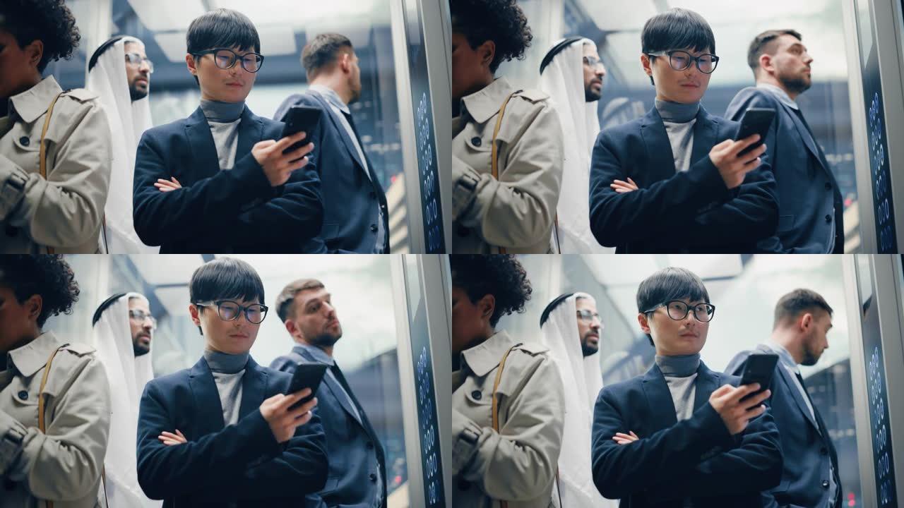 四个不同的多民族国际人士乘坐玻璃电梯在现代商务中心办公。专注于在电梯中使用智能手机的年轻时尚日本商人