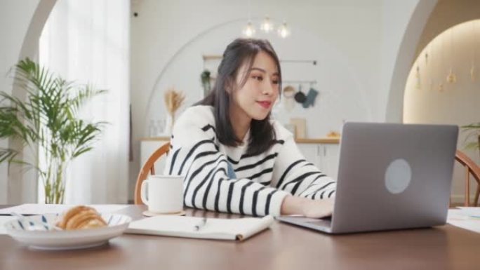 年轻的亚洲女性自由职业者专注于笔记本电脑的演示工作，以及与公司在家里厨房的远程会议的财务项目。女学生