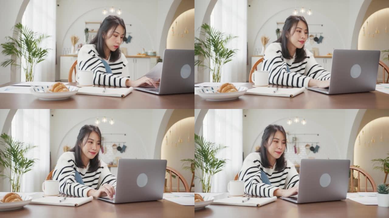 年轻的亚洲女性自由职业者专注于笔记本电脑的演示工作，以及与公司在家里厨房的远程会议的财务项目。女学生