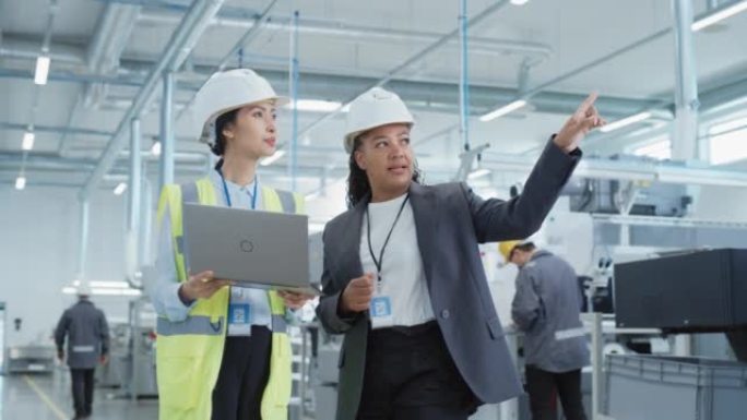 两名女重工业员工在工厂戴着安全帽。使用笔记本电脑讨论工业机械设施的工作分配。工作中的亚洲工程师和非裔