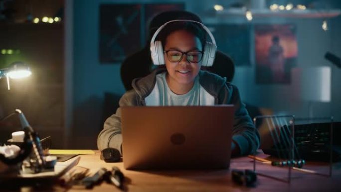 年轻的多种族女孩在家里黑暗舒适的房间里使用笔记本电脑并戴上耳机。她正在网上浏览教育研究。学习科学学校