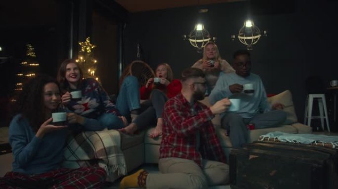 新年电影之夜。快乐的年轻多民族朋友看电视，一起在家庭派对慢动作中享受热饮。