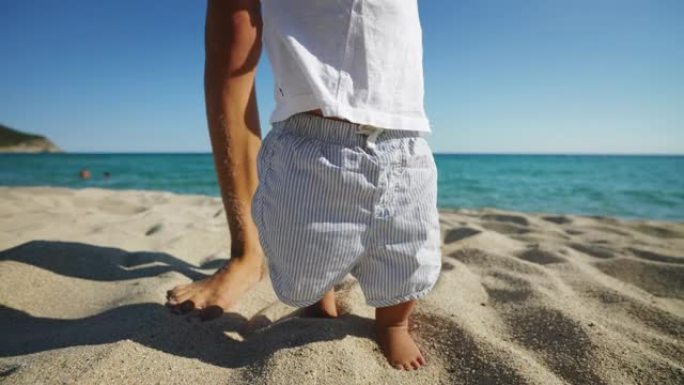 一位年轻母亲的真实特写镜头正在帮助她蹒跚学步的男婴迈出第一步，并在阳光明媚的日子里教她在海边的沙滩上