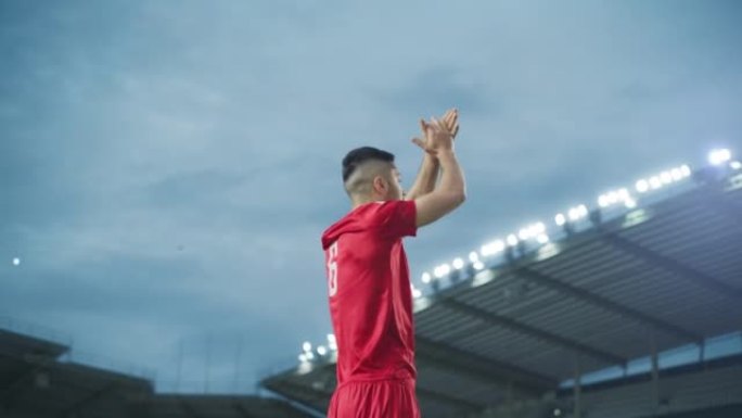 足球比赛冠军: 英俊的红队足球运动员的肖像站着，摆姿势，微笑，举手欢呼。职业足球运动员，庆祝赢得杯赛