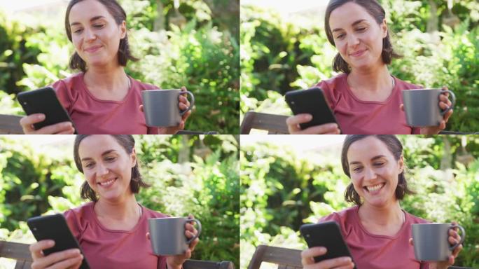 微笑的白人妇女使用智能手机，坐在阳光明媚的花园里拿着一杯茶