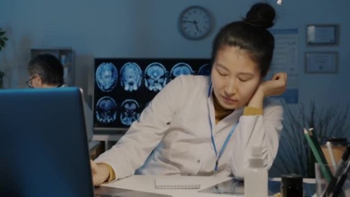 精疲力竭的亚洲医生打盹，然后晚上在黑暗的办公室里用笔记本电脑打字