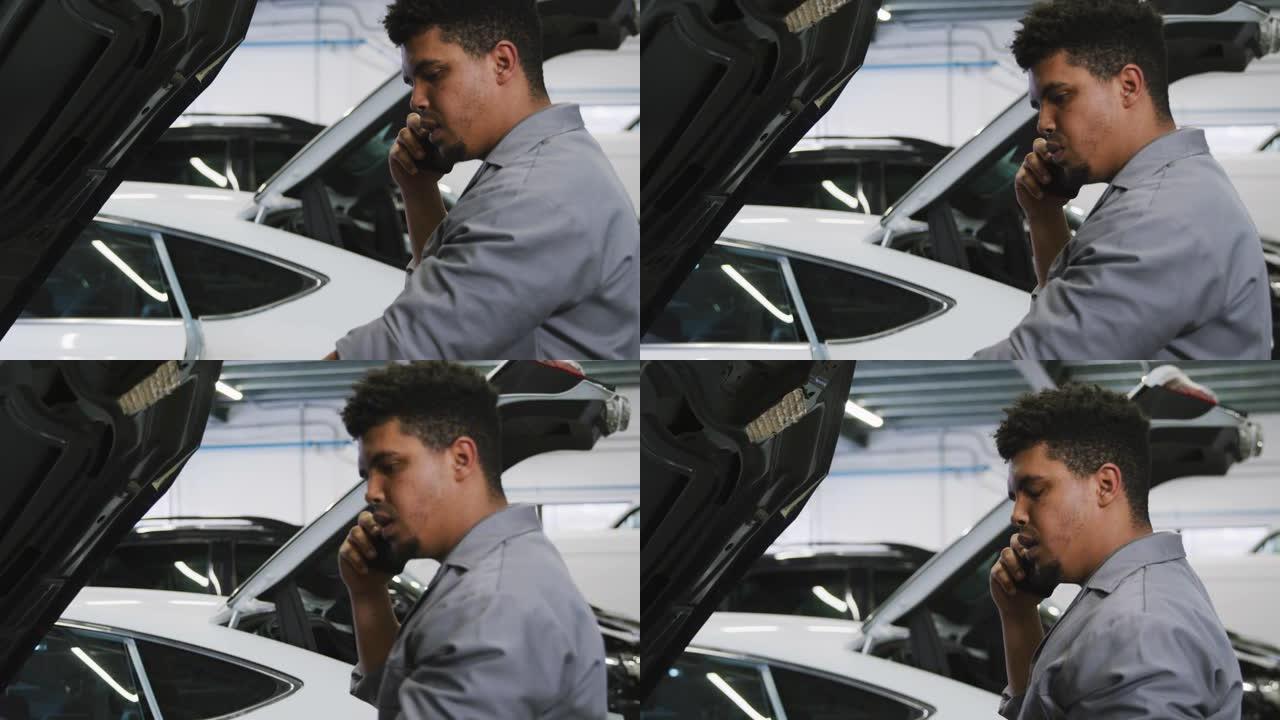 非裔美国男性汽车修理工看着打开的汽车发动机，用智能手机聊天