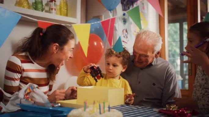 庆祝一个可爱的小男孩生日的幸福家庭肖像: 男性蹒跚学步的孩子打开礼物并拿出卡车玩具，而母亲，祖父和姐