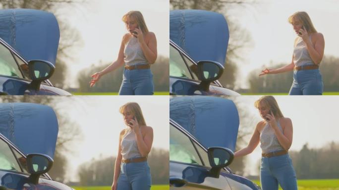 在乡间小路上沮丧的女人站在抛锚的汽车旁边，引擎盖朝上，用手机呼救-慢动作拍摄