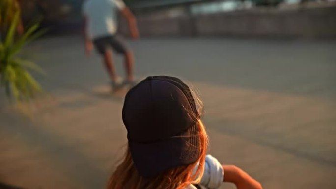 日落时，一头棕色头发的年轻女子坐在木板路上的长凳上，看着滑板手