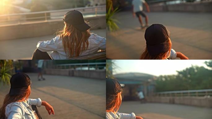 日落时，一头棕色头发的年轻女子坐在木板路上的长凳上，看着滑板手