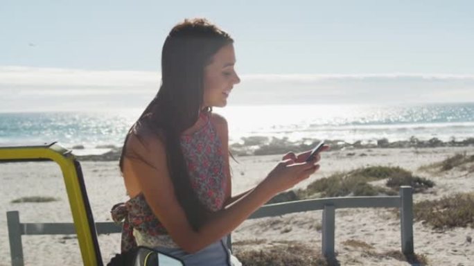 快乐的白人妇女坐在海边的沙滩越野车上，用智能手机聊天