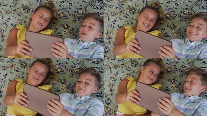 快乐的高加索兄弟姐妹在家，躺在地板上一起使用平板电脑，微笑着
