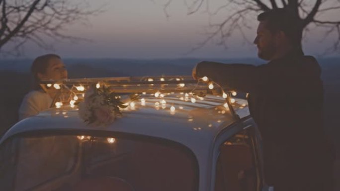 可爱的新娘和新郎靠在装饰好的汽车上，晚上互相看着对方