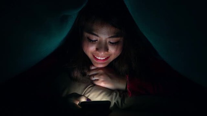 一个年轻女孩或青少年在晚上用手机和某人说话。通过她房间里的屏幕代表了现代技术在日常生活中的使用。