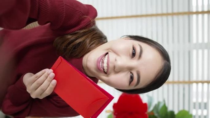 穿着红色中国新年庆祝服装的迷人年轻亚洲女士的垂直镜头快乐微笑交谈，看着相机手机拿着并展示红色零花钱或