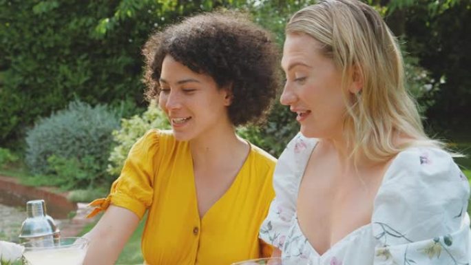 三个女性朋友坐在户外的夏季花园里，在家吃健康的饭菜，喝鸡尾酒 -- 慢动作拍摄