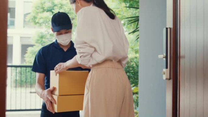 年轻的亚洲送货员戴着蓝色海军制服的口罩携带箱盒敲门家庭在线购物纸板箱给门前的女人女士在家里用智能手机