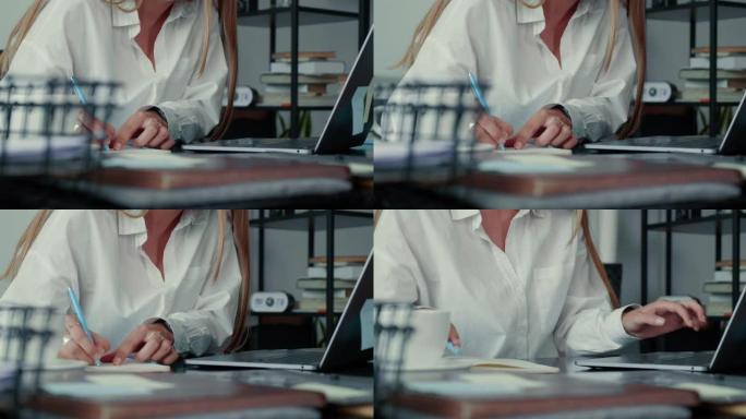特写快乐优雅的金发女商人穿着白衬衫在纸上做笔记，在现代办公桌上用笔记本电脑。