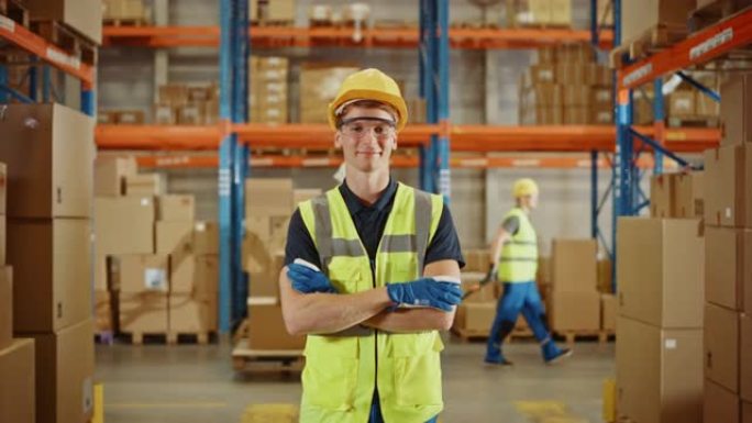 英俊的微笑工人戴着安全帽，双臂交叉站在装满纸箱的货架的零售仓库中。物流、配送和配送中心的专业工作