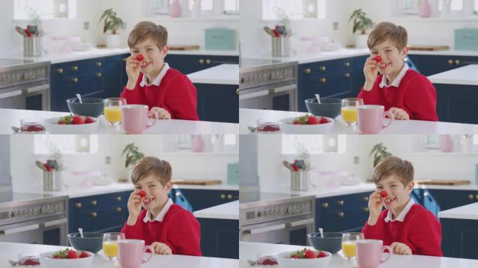 笑着的男孩穿着校服在厨房里吃早餐时把草莓放在鼻子上