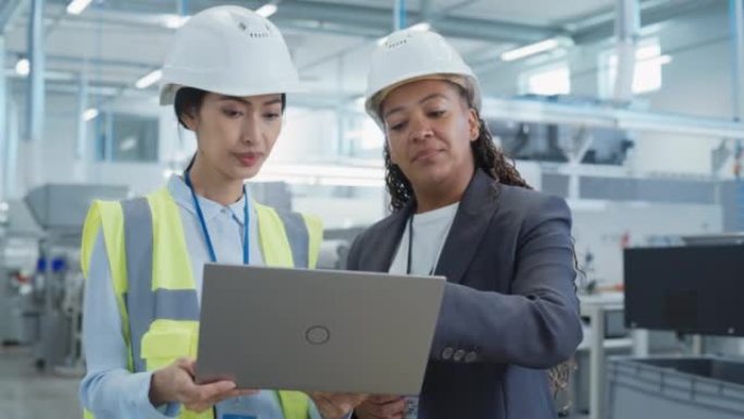 两名戴着安全帽的女员工在工厂的肖像。使用笔记本电脑讨论工业机械设施的工作分配。工作中的亚洲工程师和非