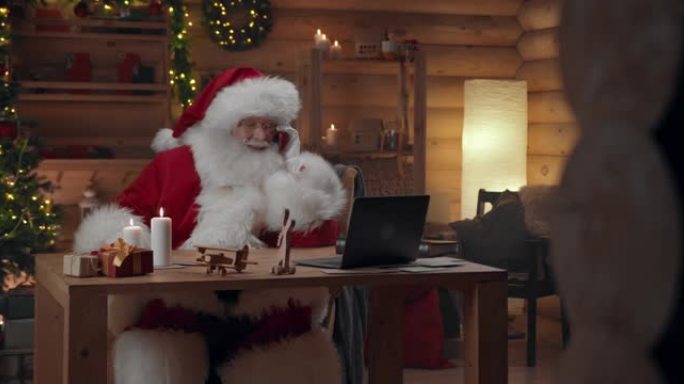 圣诞老人正在用红色智能手机聊天，坐在办公室的桌子上