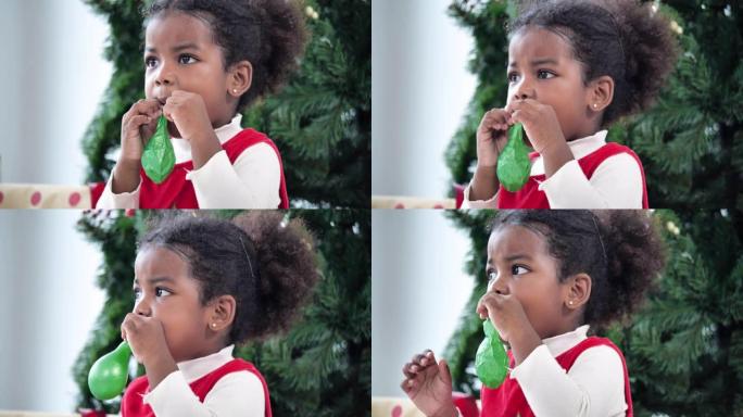 非洲民族的小女孩年龄3岁的威灵红色衣服在圣诞节在家吹绿色气球快乐又有趣。亲密的圣诞节概念。
