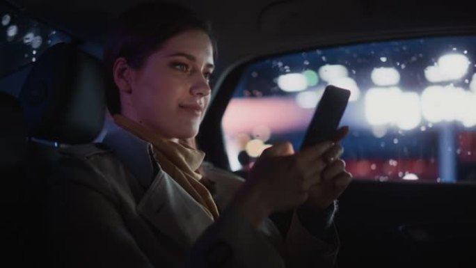时尚的女性晚上坐出租车后座回家。美丽的女人乘客使用智能手机，在城市街道上开着霓虹灯的汽车时看着窗外。