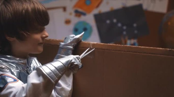 有趣的DIY概念。穿着太空服的可爱的小男孩看着相机，用剪刀在他的房间里剪大纸板。