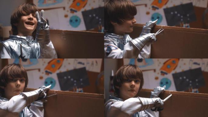 有趣的DIY概念。穿着太空服的可爱的小男孩看着相机，用剪刀在他的房间里剪大纸板。