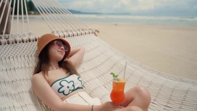 快乐的年轻女子，喝鸡尾酒，在海滩的吊床上放松。