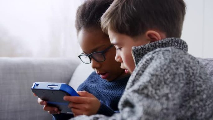 两个小男孩坐在家里的沙发上，一起在家里的手持设备上玩游戏