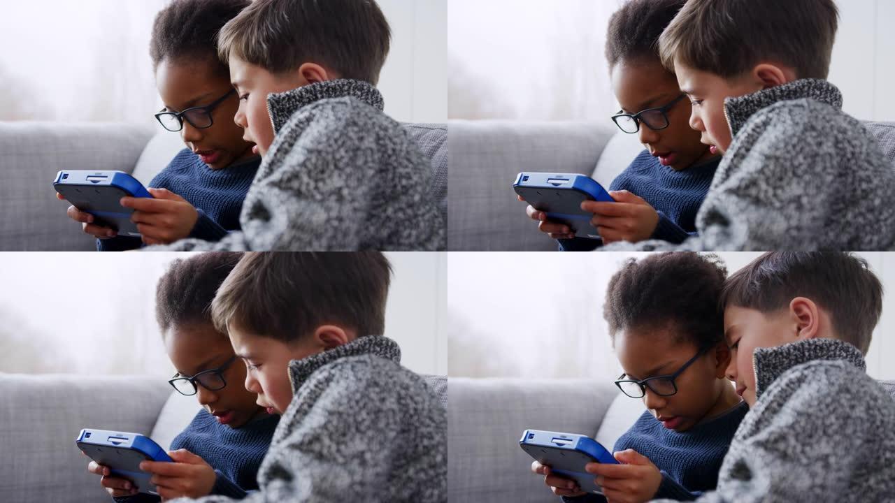 两个小男孩坐在家里的沙发上，一起在家里的手持设备上玩游戏