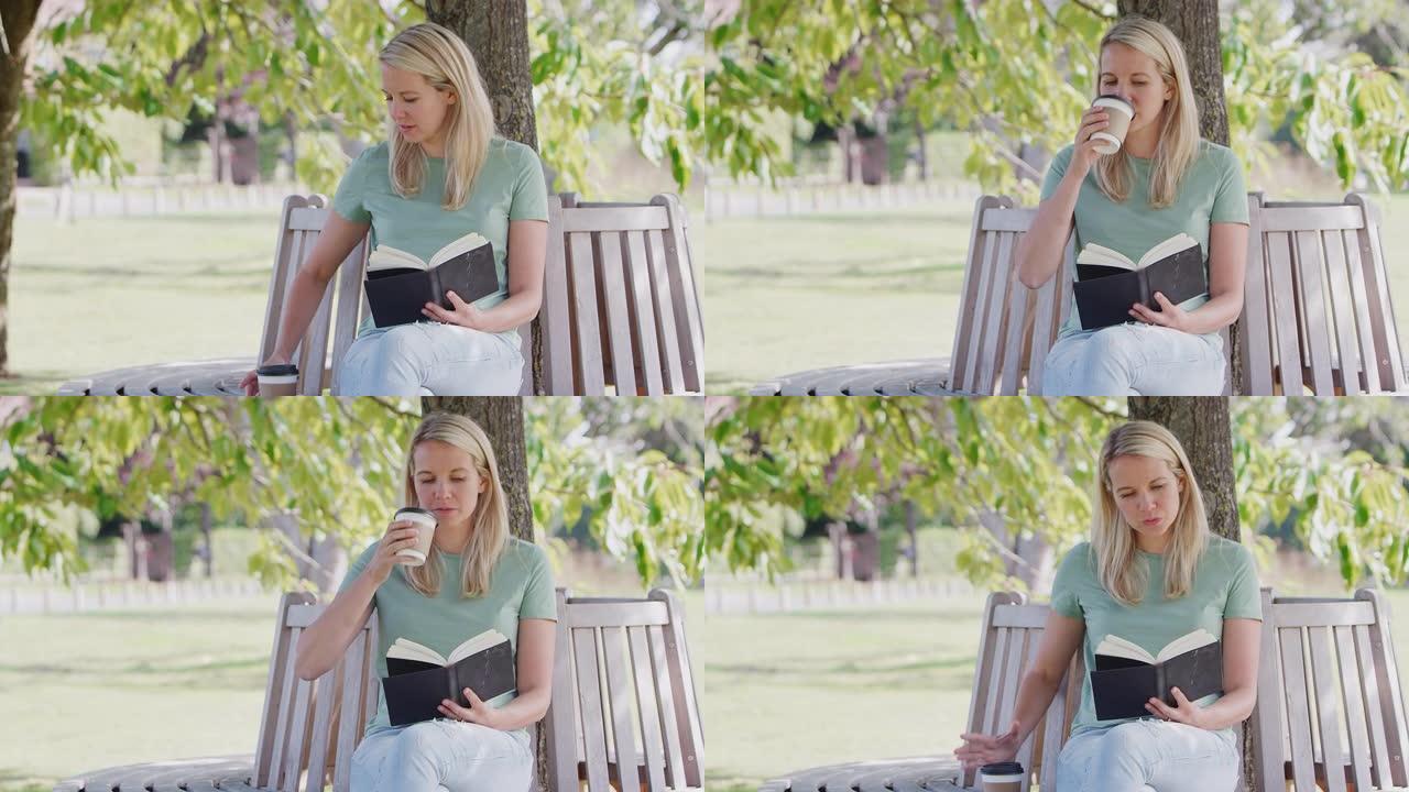 女人坐在公园的长椅上，在树下看书，喝外卖咖啡