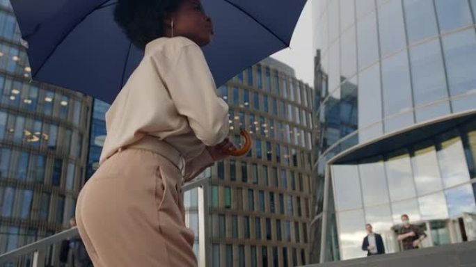 一位穿着米色上衣的年轻黑皮肤妇女走上楼梯，在下雨天用两只手拿着深蓝色的雨伞