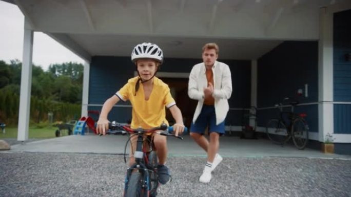 开朗的父亲在教儿子在他们美丽的住宅区的前院骑自行车。男孩戴着头盔，勇敢地骑着自行车平衡自己。手持镜头