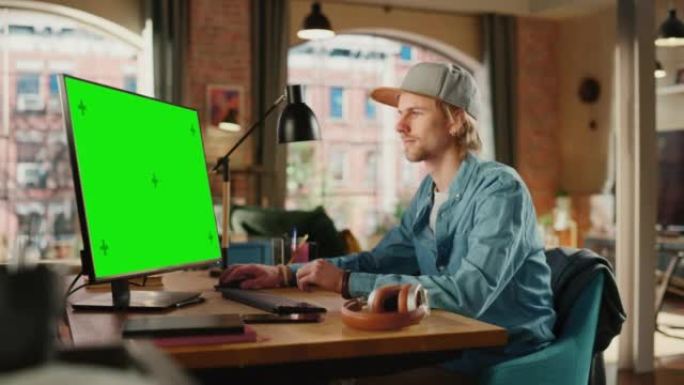 年轻英俊的男人在家工作，在带有绿屏模拟显示的台式计算机上。创意男性查看社交媒体，浏览互联网。从大窗户