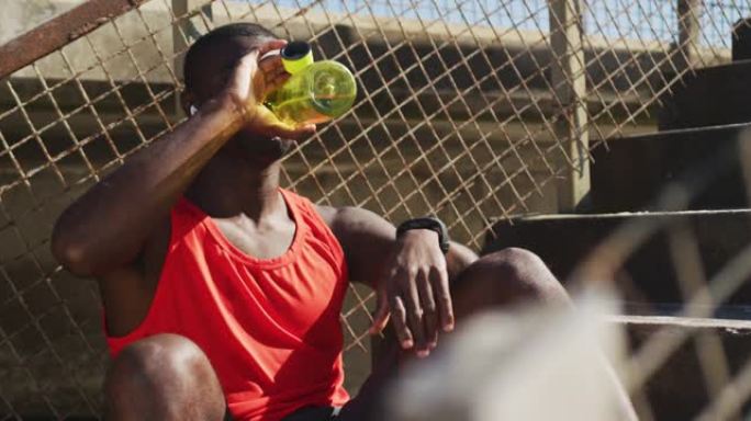 疲惫的非洲裔美国男子坐着，用水瓶喝水，在户外锻炼休息