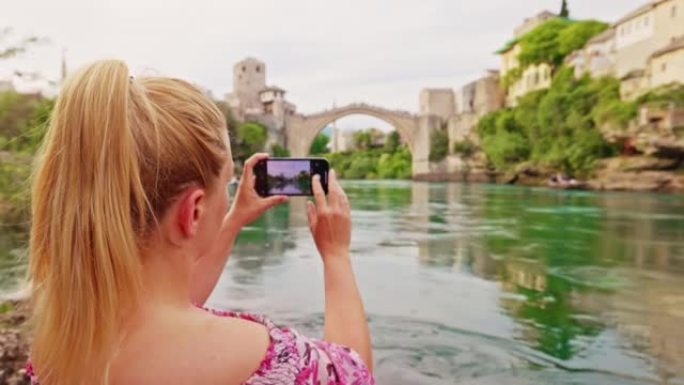 金发碧眼，粉红色连衣裙的特写女人坐在neretva河上，用智能手机拍摄了Stari most，Mos