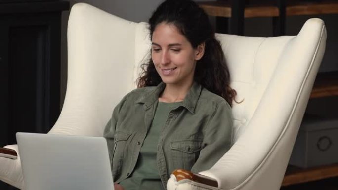 快乐的年轻女子坐在舒适的扶手椅上，使用电脑应用程序。