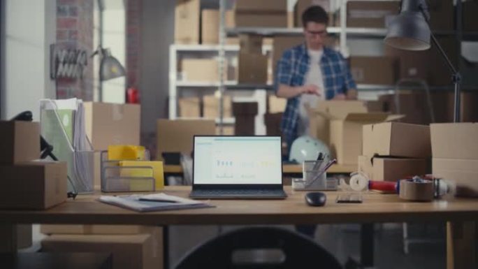 笔记本电脑站在桌子上，显示屏上实时统计销售数据。小型企业仓库，工人在后台行走。带纸箱的桌子。
