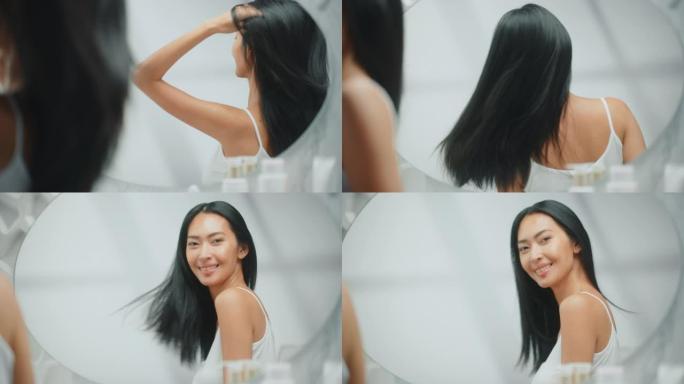 美丽的亚洲女人围着她郁郁葱葱的黑发飞舞。年轻的成年女性在浴室镜子里欣赏自己。幸福、自然美、有机护肤产