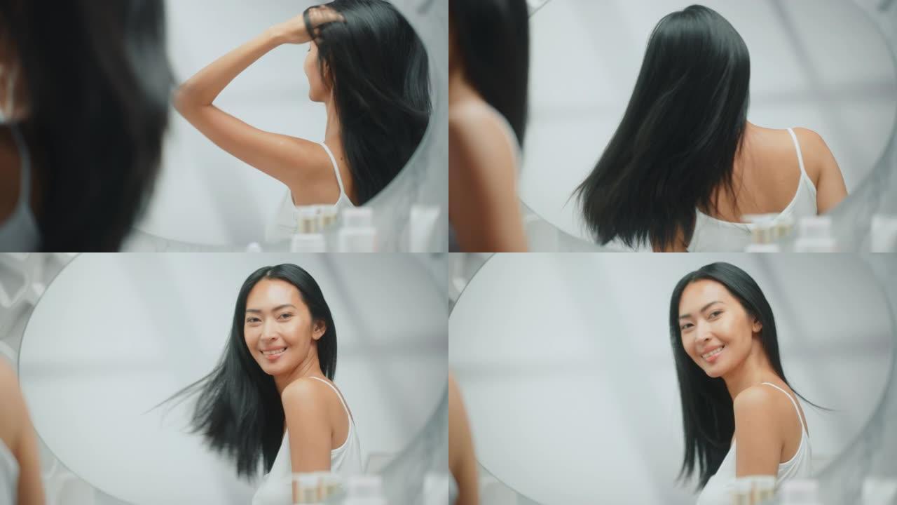 美丽的亚洲女人围着她郁郁葱葱的黑发飞舞。年轻的成年女性在浴室镜子里欣赏自己。幸福、自然美、有机护肤产