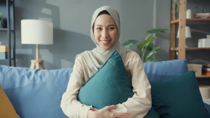 迷人开朗的年轻亚洲穆斯林美女戴着头巾，随意坐在沙发上微笑，在家舒适的客厅看着相机。女孩伊斯兰社会距离