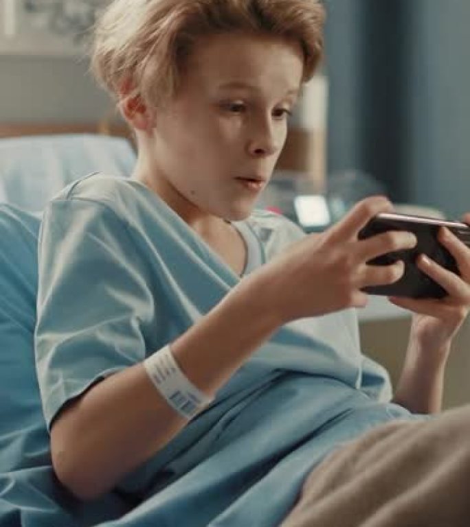 垂直屏幕。医院病房: 英俊的小男孩躺在床上休息，使用智能手机，用手机在互联网上玩在线视频游戏。生病后